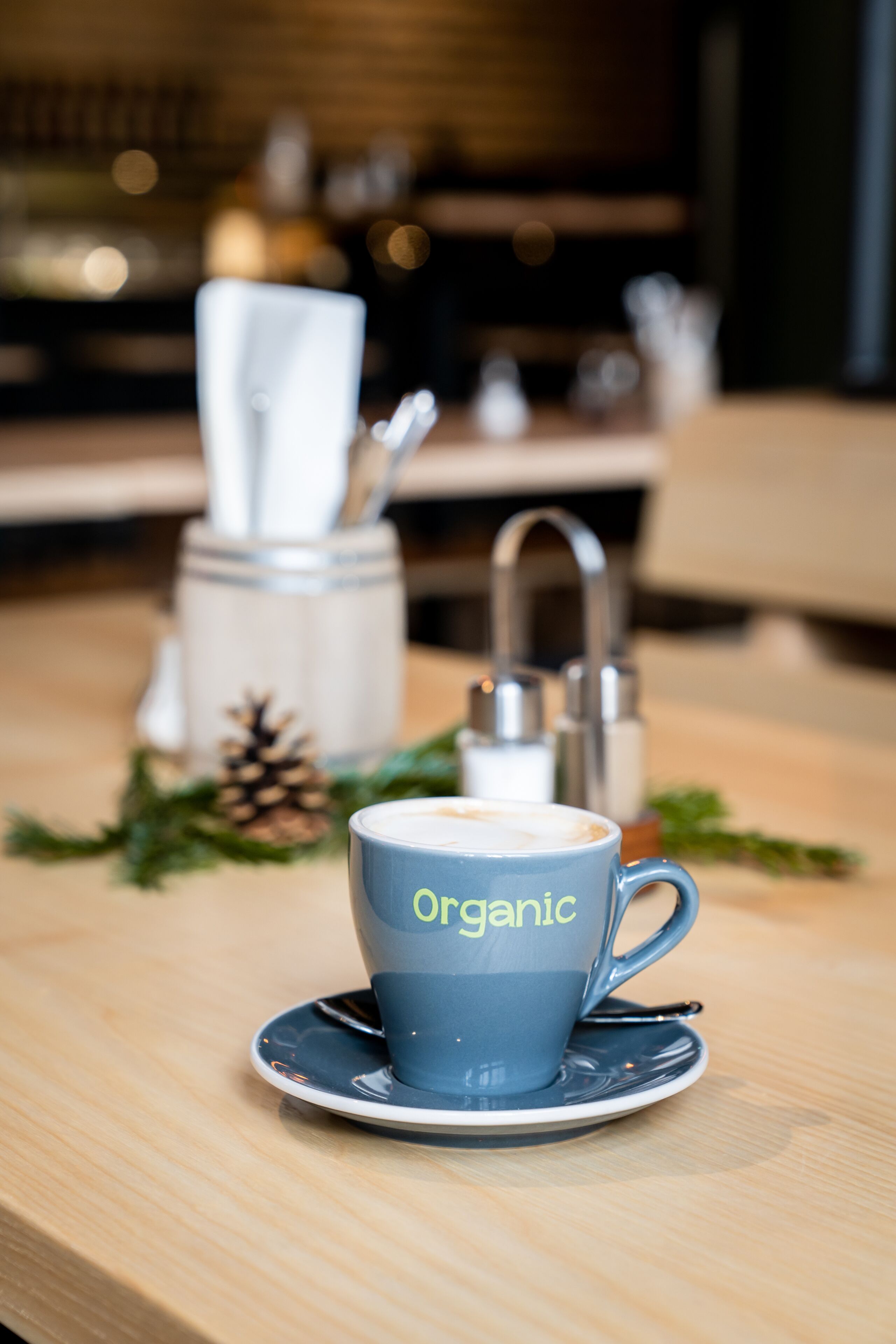 A coffee cup labeled "organic" on a saucer, with a blurred background featuring a coffee machine and a wooden table.