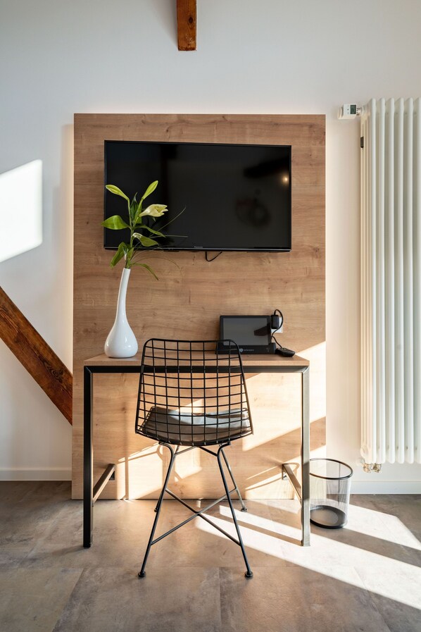 Un espacio de trabajo moderno con un escritorio de madera, una silla negra, un jarrón blanco con una planta, un televisor montado en la pared y un radiador.