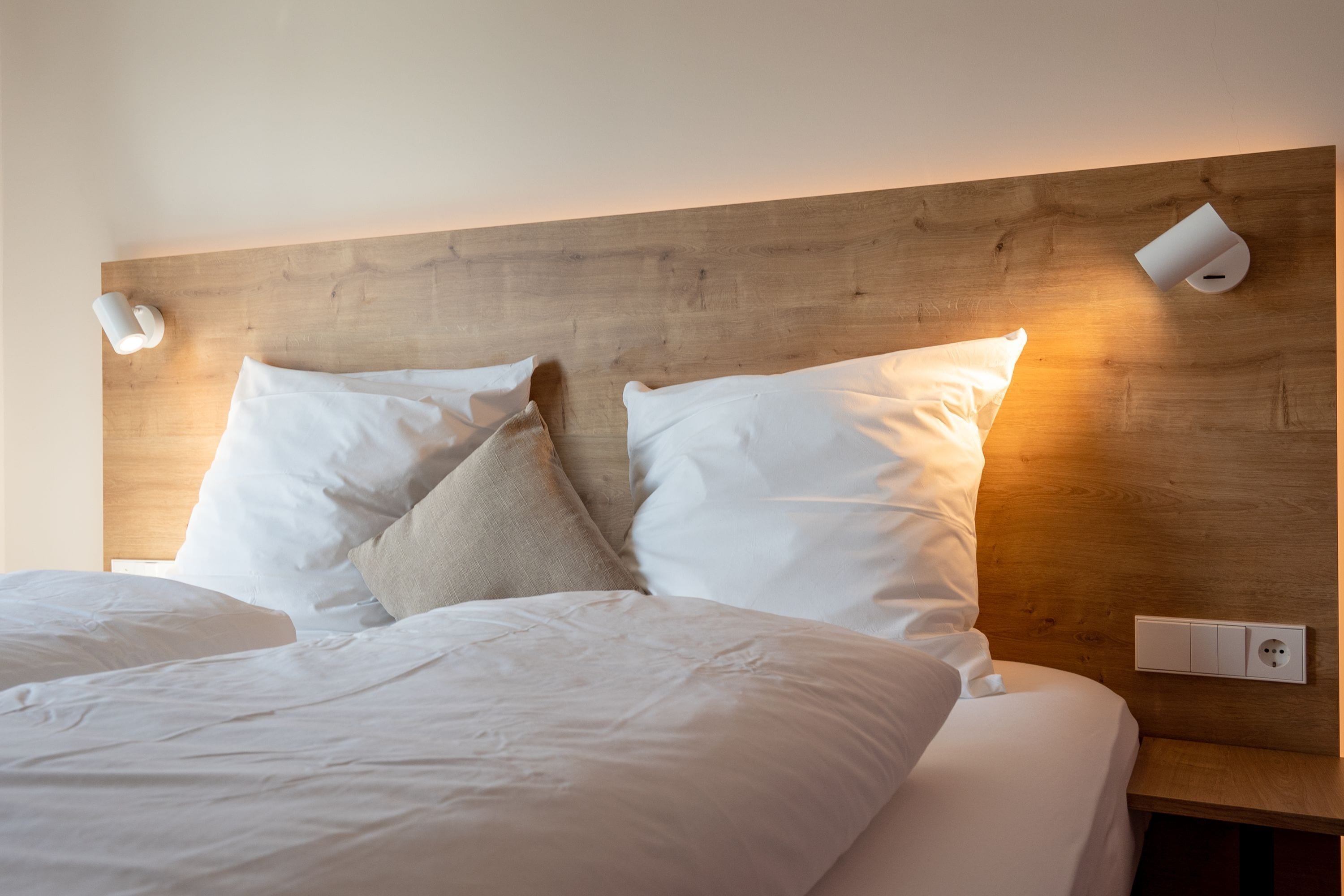 Una cama bien hecha con almohadas blancas y beige, un cabecero de madera y dos lámparas fijadas a la pared.