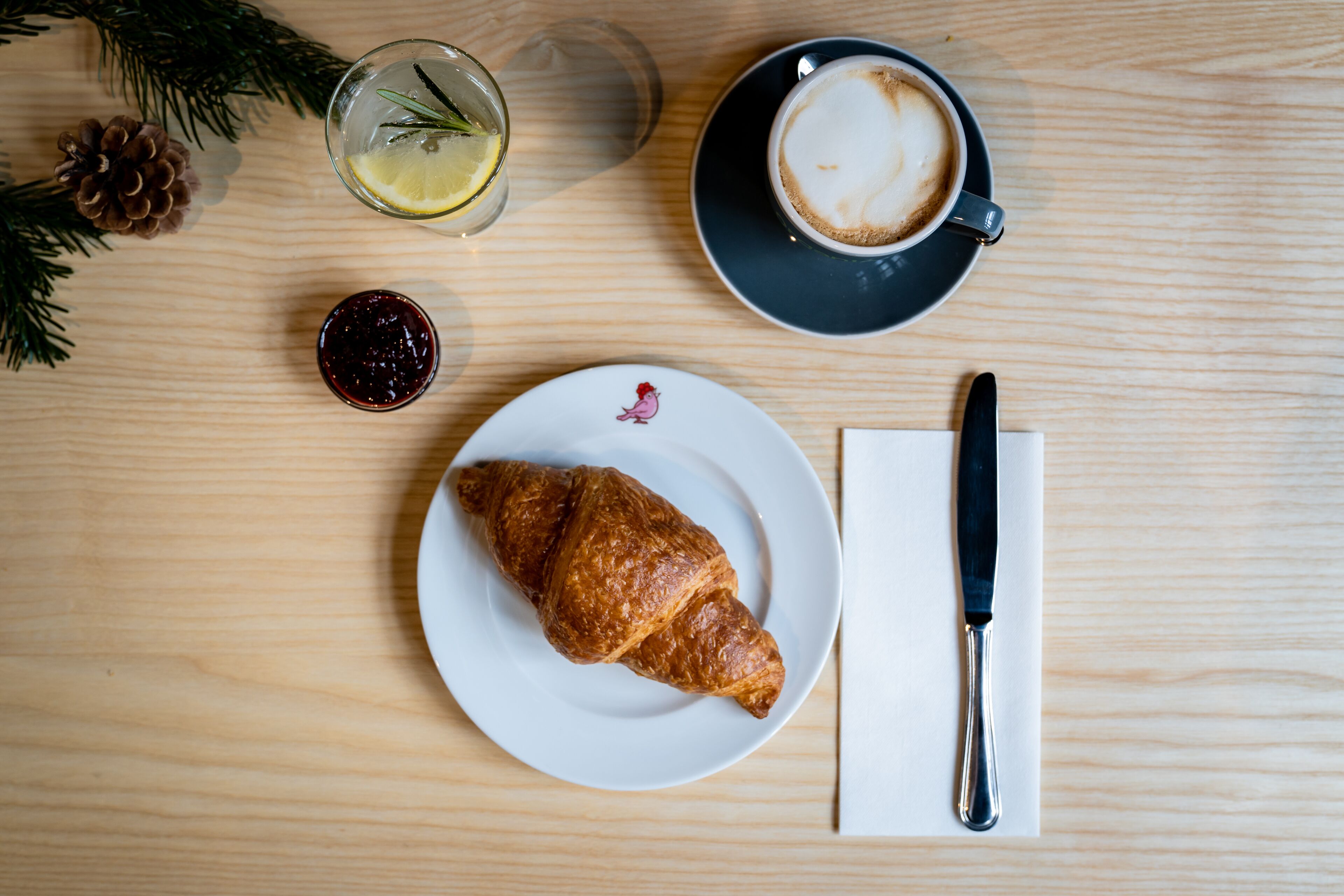 Une table en bois avec un croissant, une tasse de café, un verre d’eau au citron et un petit pot de confiture.