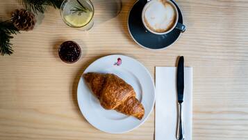 Une table en bois avec un croissant, une tasse de café, un verre d’eau au citron et un petit pot de confiture.