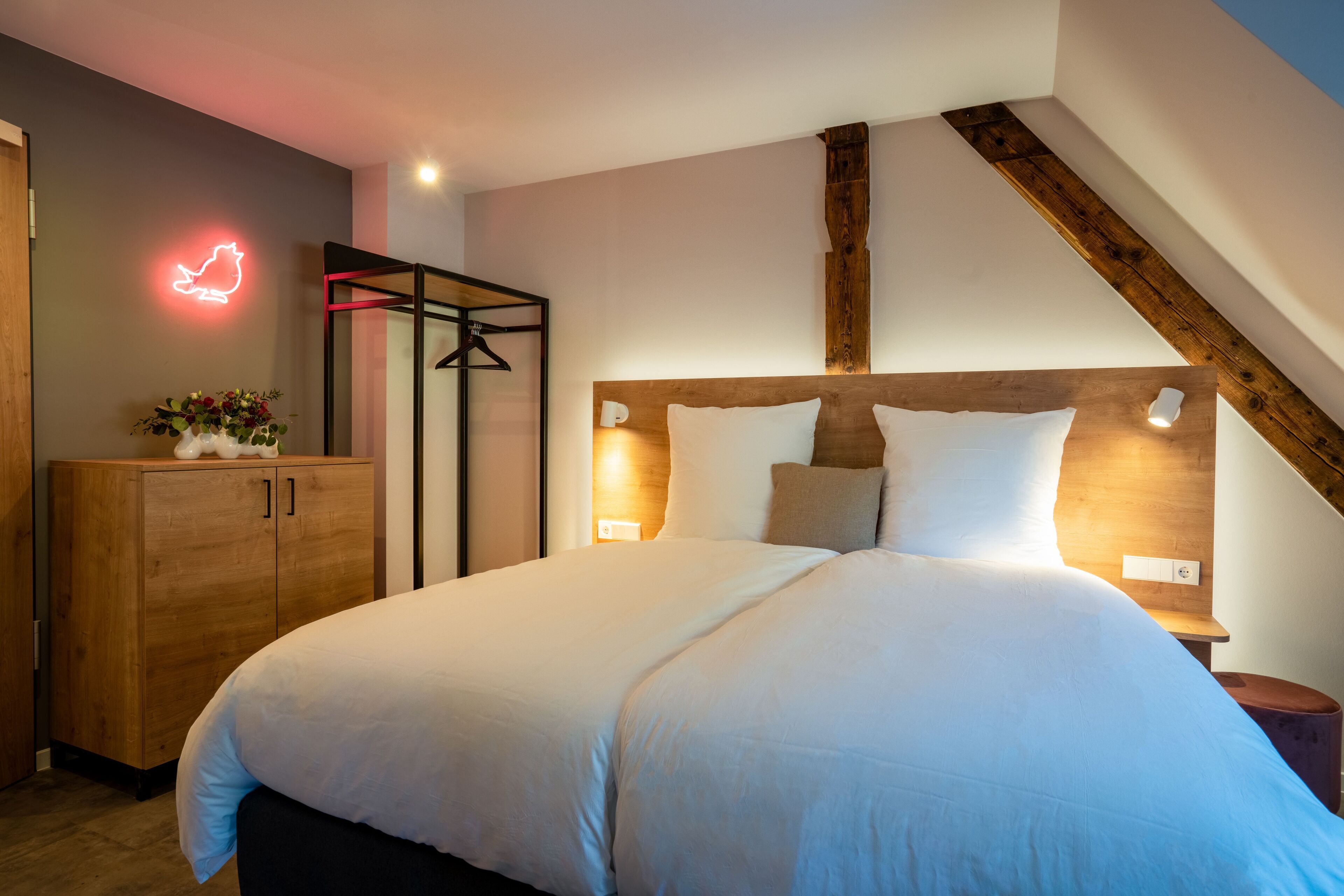 Ein modernes Schlafzimmer mit einem hölzernen Kopfteil, einem Bett mit weißer Bettwäsche, einem Holzschrank und einem leuchtenden Katzen-Schild an der Wand.