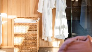 Une pièce aux murs lambrissés, avec un plafond en pente, un panier à linge en osier et deux robes blanches suspendues à un porte-vêtements.
