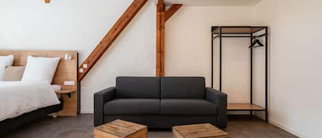 Una habitación moderna con cama, sofá gris oscuro, cajas de madera, armario metálico y una escalera de madera.