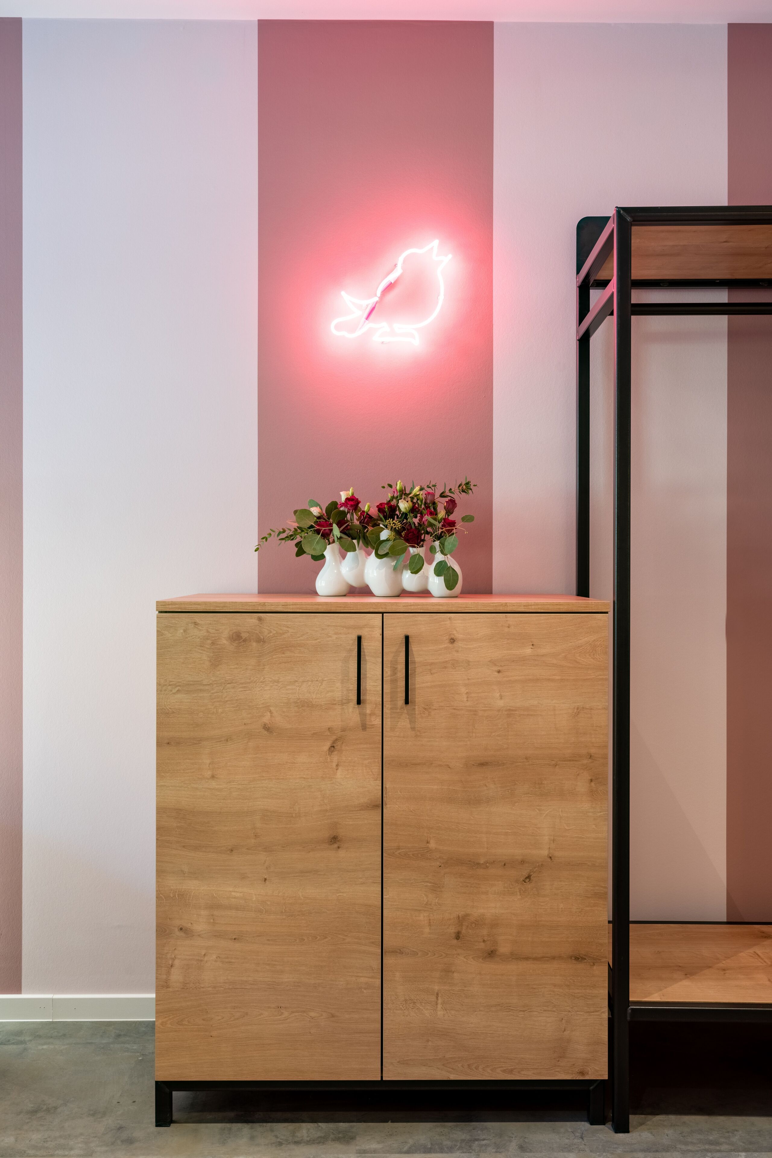 A wooden cabinet with a neon sign, a small table with flowers, and a bunk bed against a wall with vertical stripes.