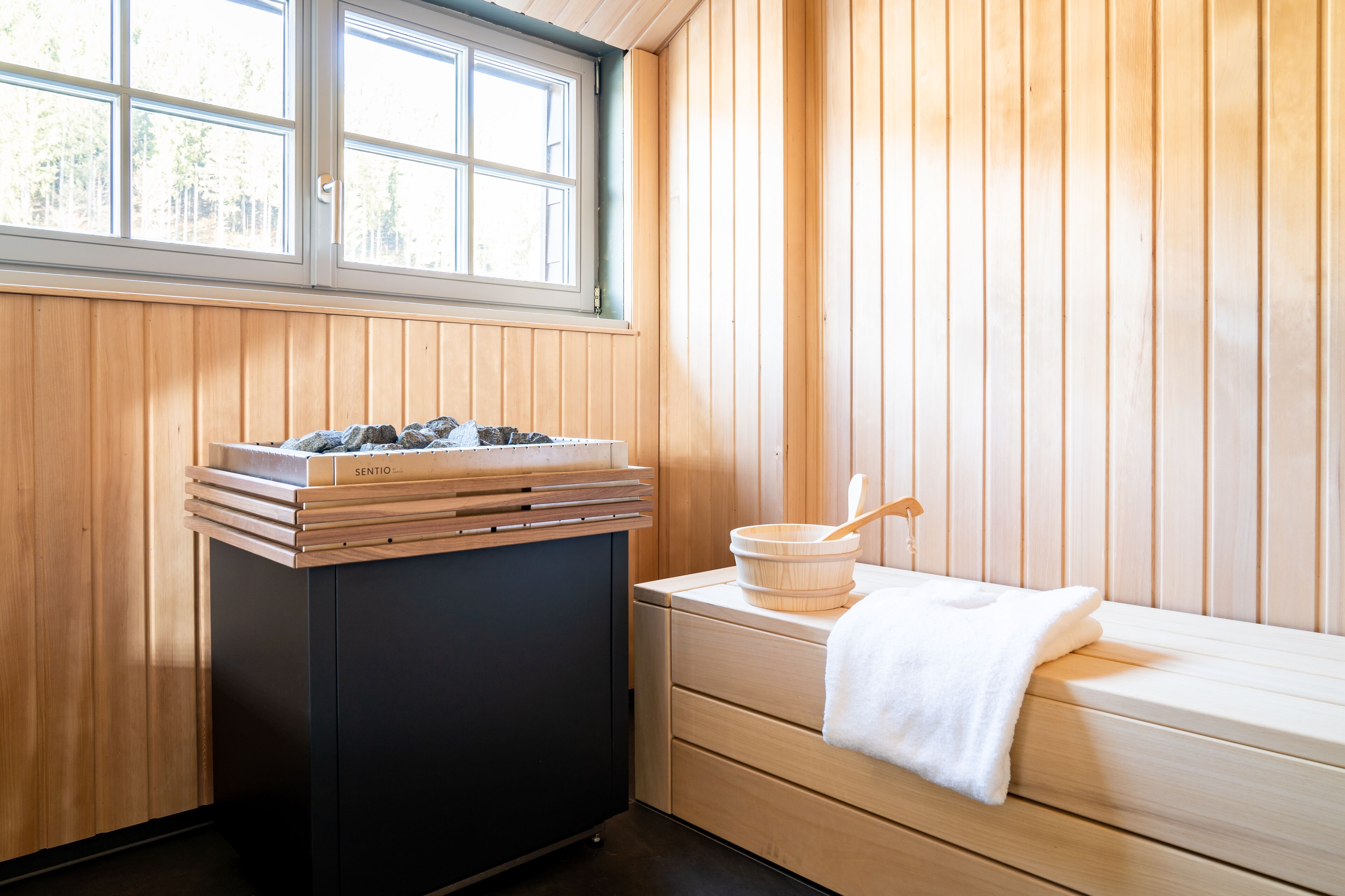 A sauna room with wooden walls, a window, a massage table, and a wooden cabinet with towels and a wooden bowl.