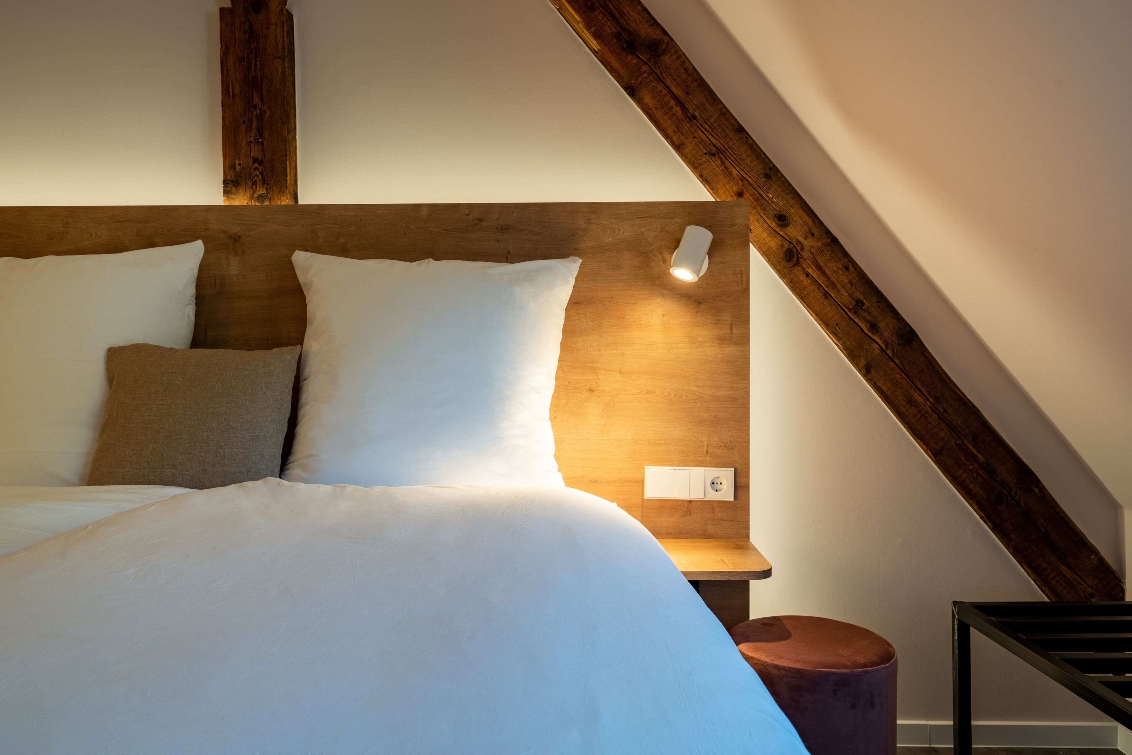 A neatly made bed with white linens and a wooden headboard, a small bedside table, and a wall socket.