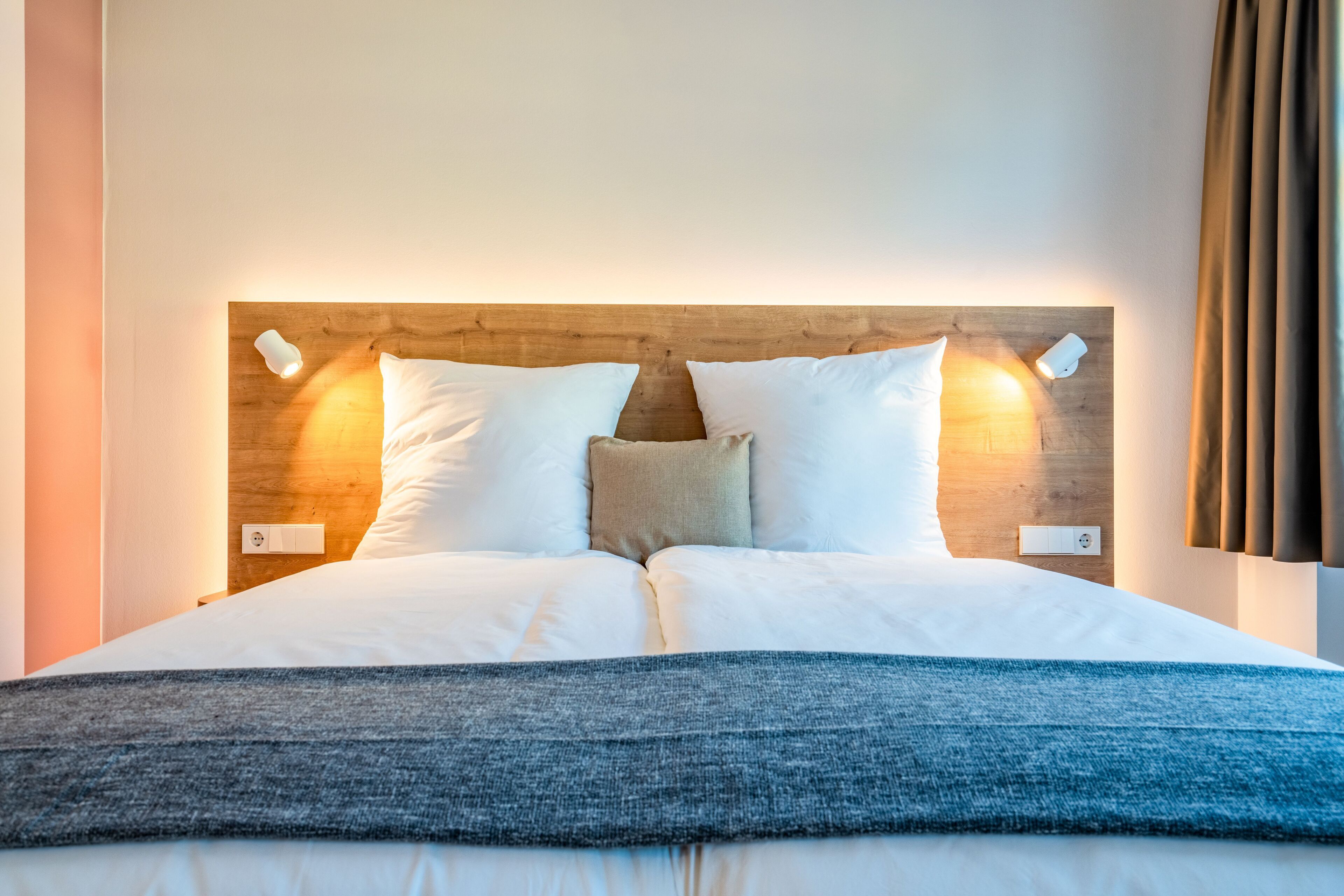 A neatly made bed with white bedding and a blue throw blanket, a wooden headboard with wall-mounted lights, and a power outlet on the wall.