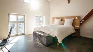 Une chambre à coucher moderne avec un grand lit, un banc et une vue sur la ville grâce à de grandes fenêtres.