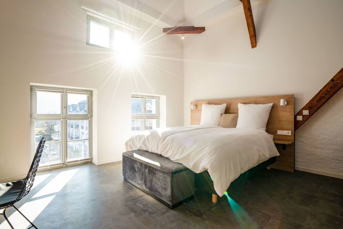 A modern bedroom with a large bed, a bench, and a view of the city through large windows.