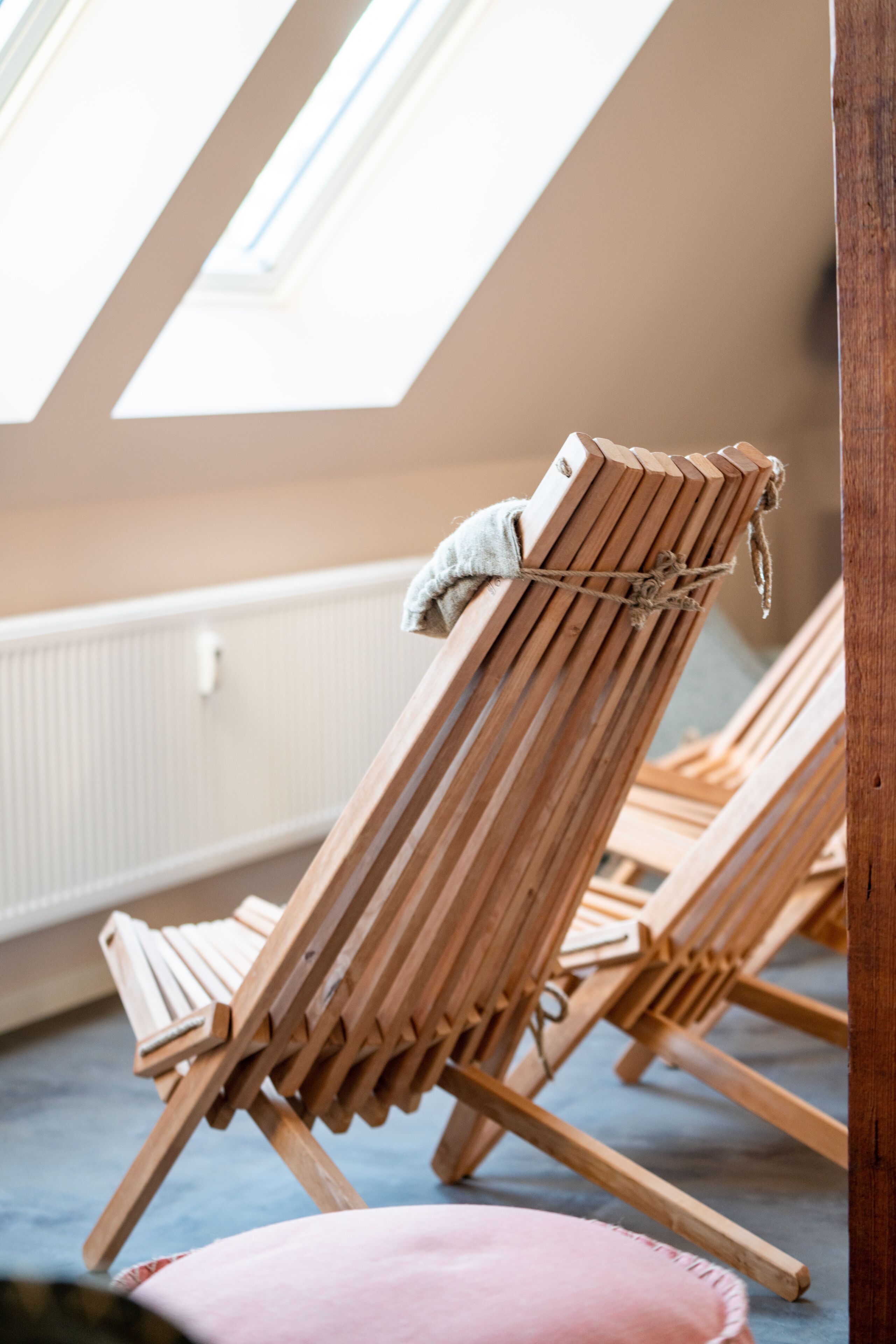 Ein Zimmer mit schrägem Dach und Oberlichter, Holz-Loungestühle mit Kissen und ein Heizkörper an der Wand.