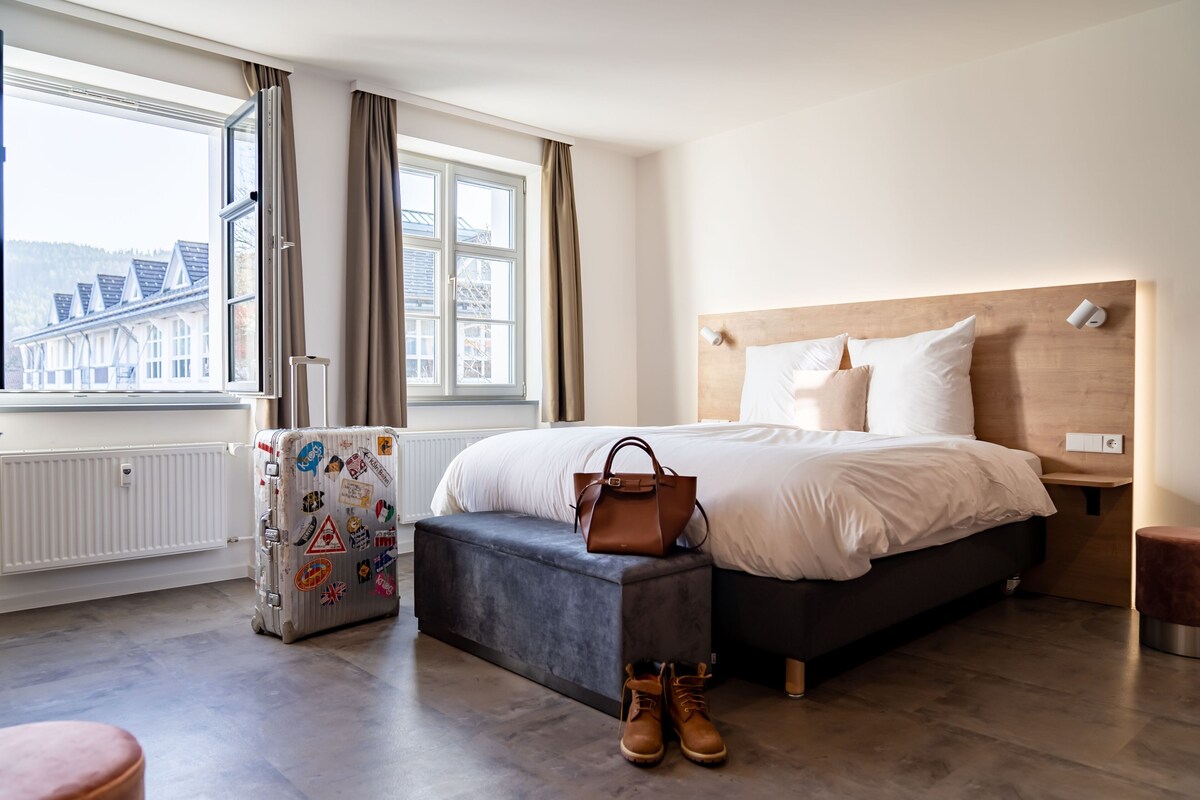 A bedroom with a bed, a suitcase, a handbag, and a window with curtains.