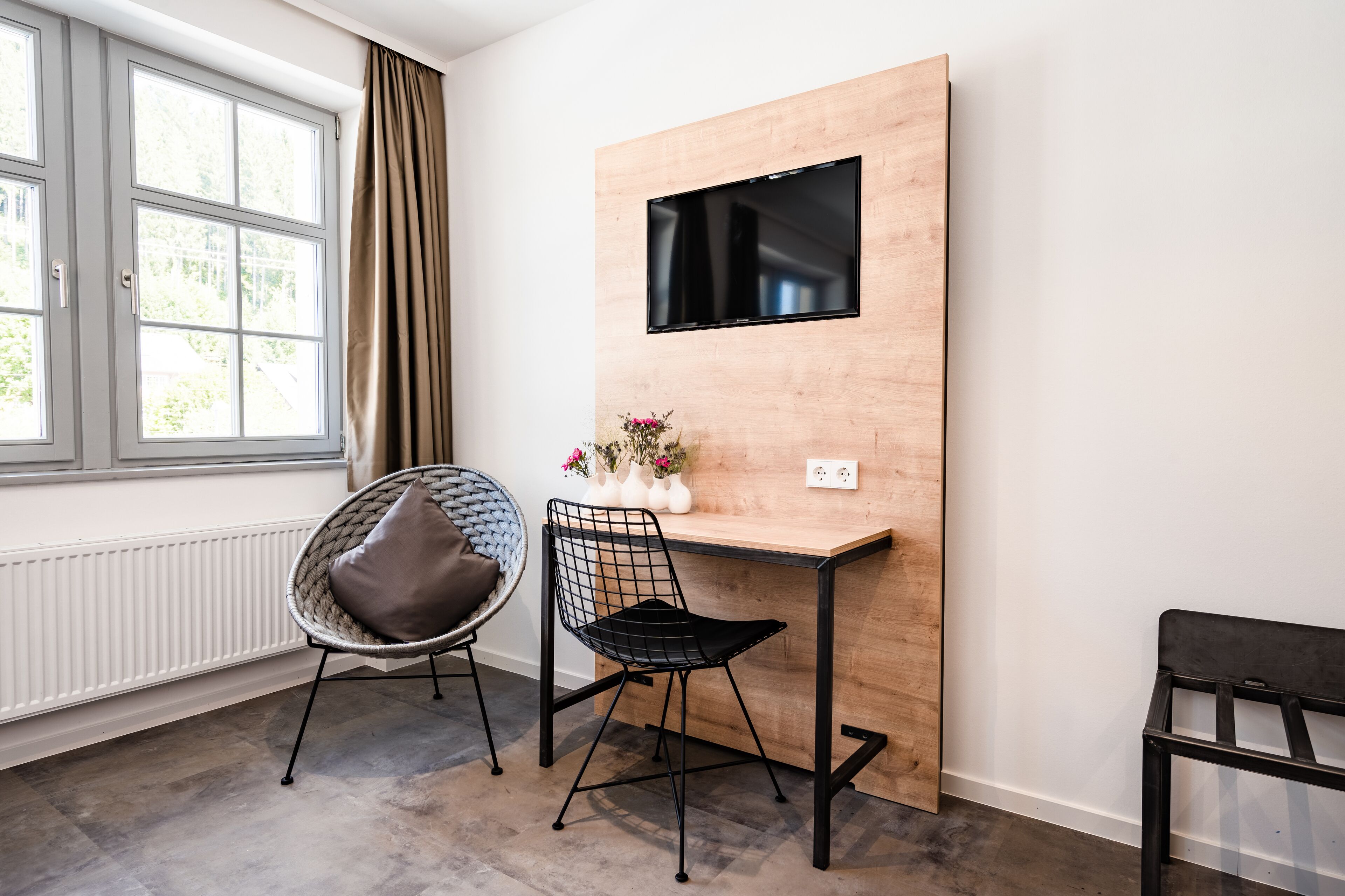 A modern room with a wooden desk, two chairs, a TV mounted on the wall, and a window with curtains.