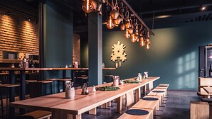 Un restaurant moderne avec de longues tables et des bancs en bois, des casseroles en cuivre suspendues et un mur végétal agrémenté d’une décoration en forme d’engrenage en bois.