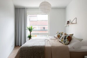 A modern bedroom with a large window, a bed with floral pillows, a hanging light fixture, and a small plant.
