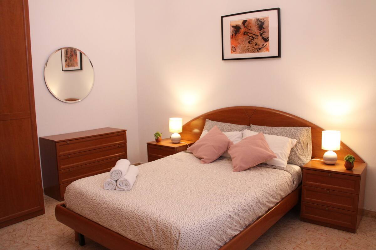 A neatly made bed with a wooden headboard, two bedside tables with lamps, a round mirror on the wall, and a framed picture above the bed.