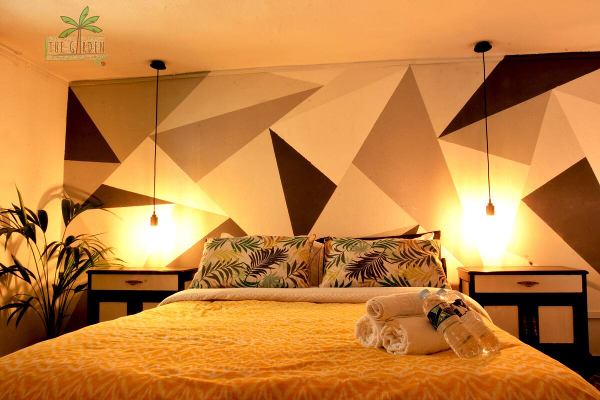 A hotel room with a geometric patterned wall, a bed with a yellow patterned bedspread, two bedside tables with lamps, and a potted plant.