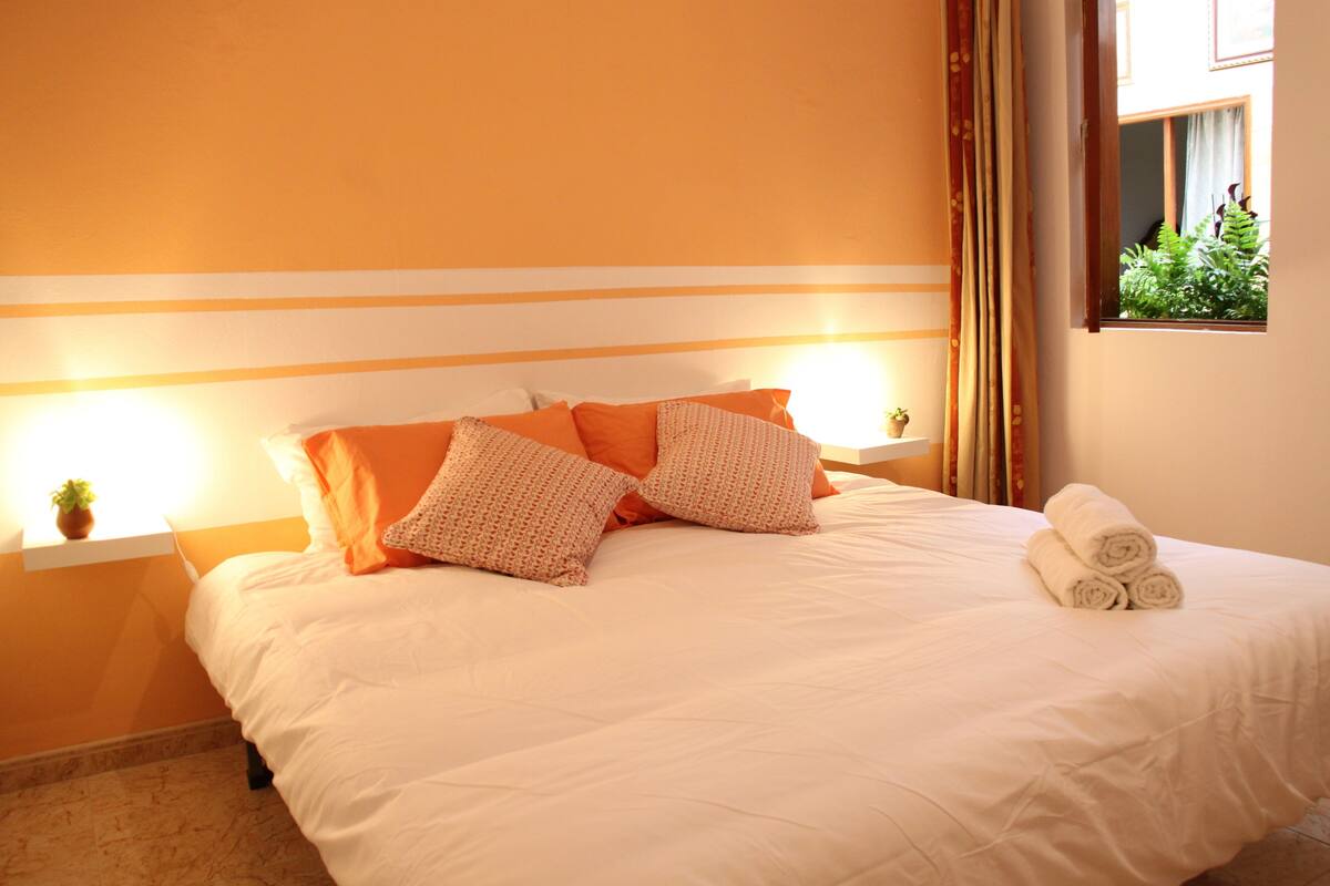 A neatly made bed with white bedding and orange pillows, a small potted plant on the nightstand, and a window with curtains.