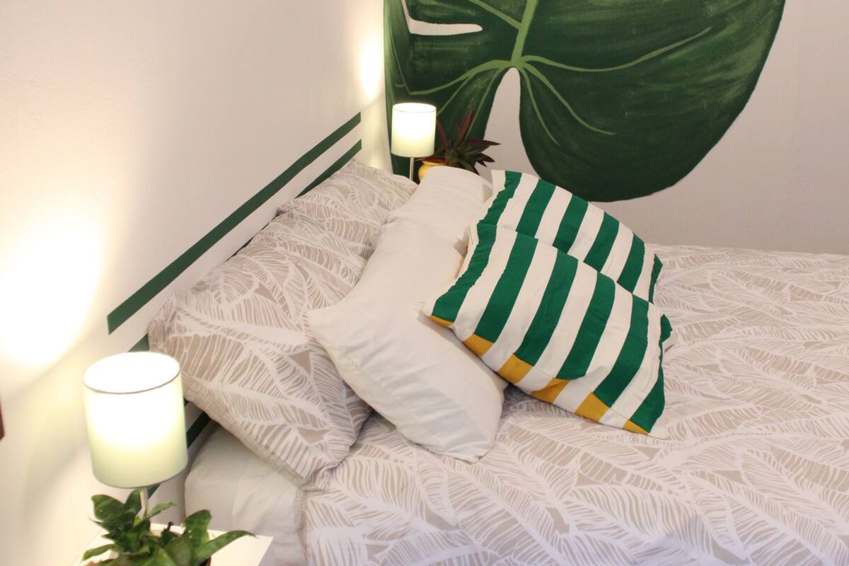 A bedroom with a large green leaf mural, a white patterned bedspread, a striped pillow, a bedside lamp, and a small potted plant.