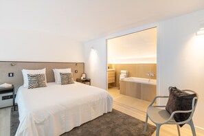 Moderni hotellihuone, jossa on suuri sänky, yöpöytä, tuoli ja avoimesta ovesta näkyvä kylpyhuone.