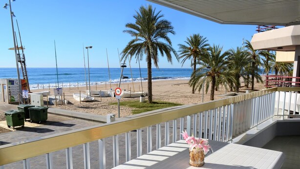 A balcony with a white table, a flower vase, and a view of the beach with palm trees and the ocean. - Sant Telm A - Calafell (Calafell)