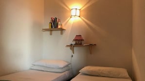 Une petite pièce avec deux lits simples, une étagère fixée au mur avec des livres et une lampe, et un luminaire fixé au mur.