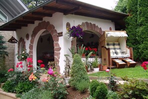 A traditional house with a stone facade, a wooden porch, and a garden with various plants and flowers.