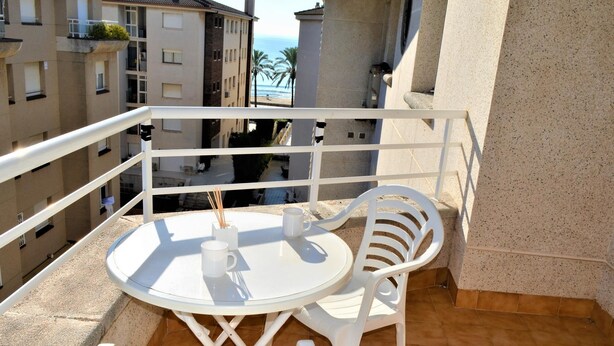 A balcony with a round table and two white chairs, overlooking a coastal area with buildings and palm trees. - Apartment Barcino - Calafell (CALAFELL)