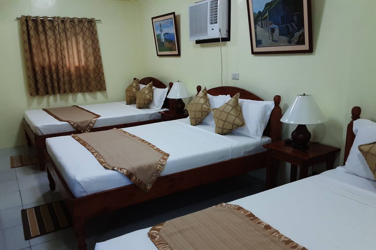 A hotel room with three wooden beds, each with white linens and brown throw blankets. There are bedside lamps, a wall-mounted air conditioner, and framed artwork on the walls.