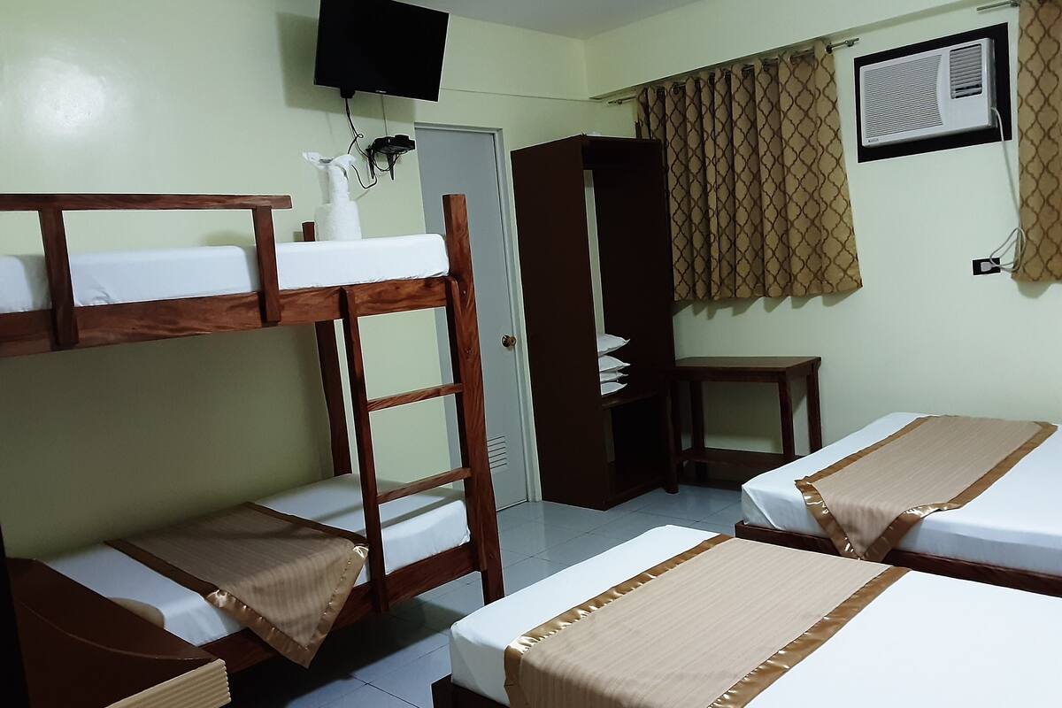 A dormitory room with bunk beds, a desk, a wardrobe, a television, and a window with curtains.