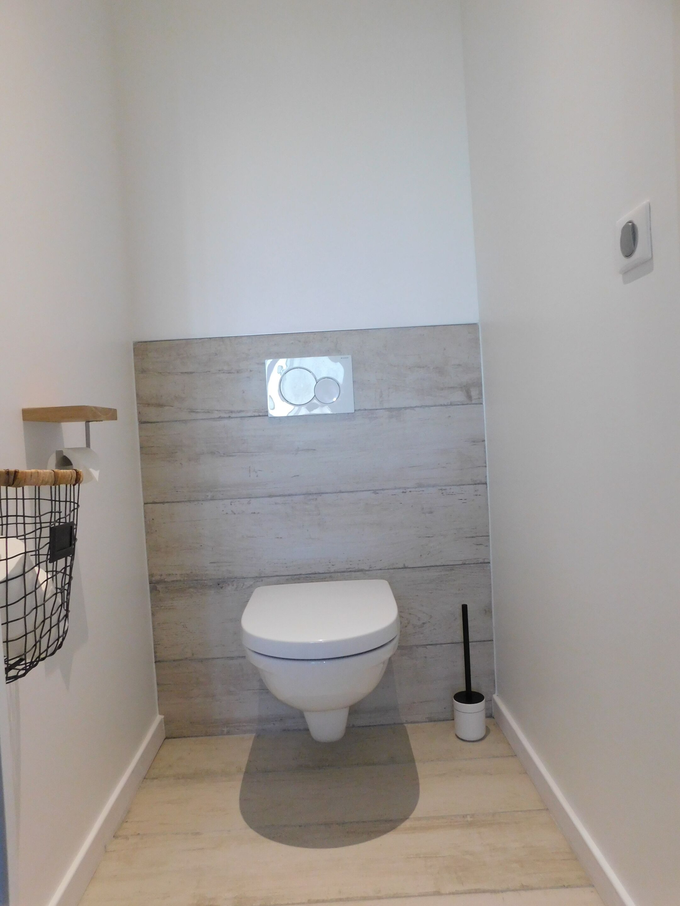 Ein kleines, modernes Badezimmer mit einer weißen Toilette, einem Holzregal und einem an der Wand befestigten Spiegel.