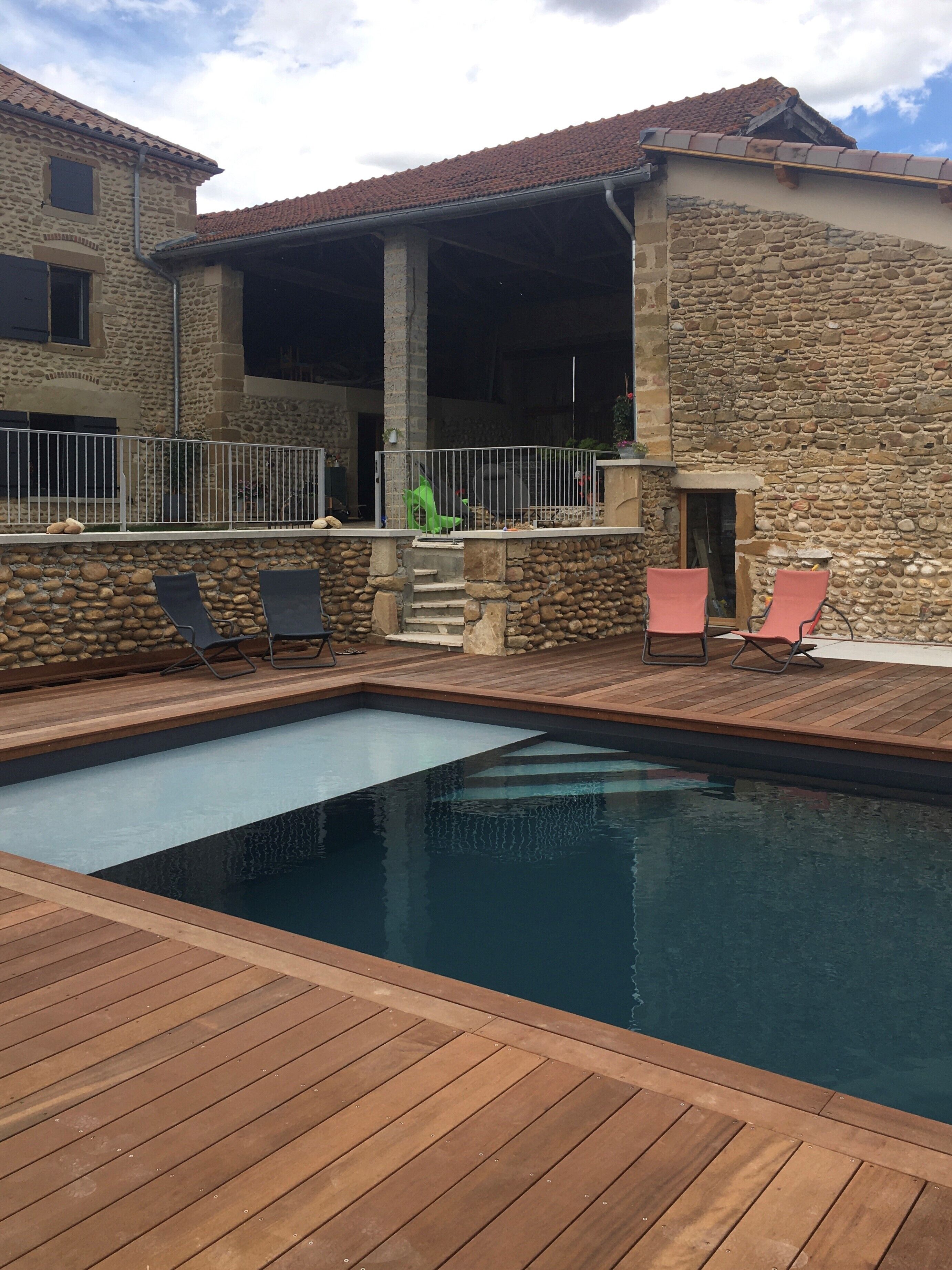 Una piscina amb zona de solàrium de fusta i gandules, envoltada per parets de pedra i un porxo cobert.