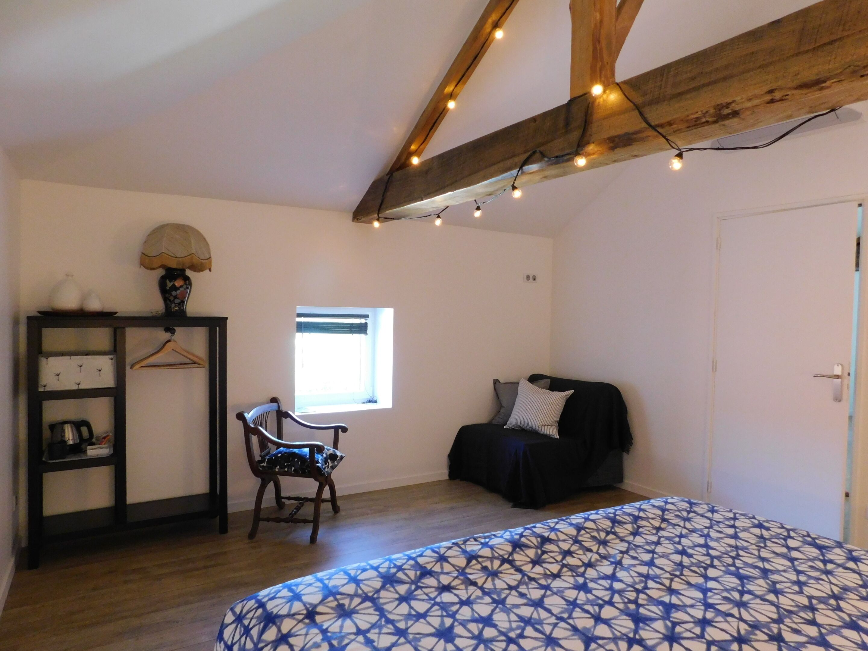 A bedroom with a bed, a chair, a small table, a shelf with decorative items, a window, and exposed wooden beams.