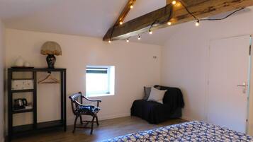 A bedroom with a bed, a chair, a small table, a shelf with decorative items, a window, and exposed wooden beams.