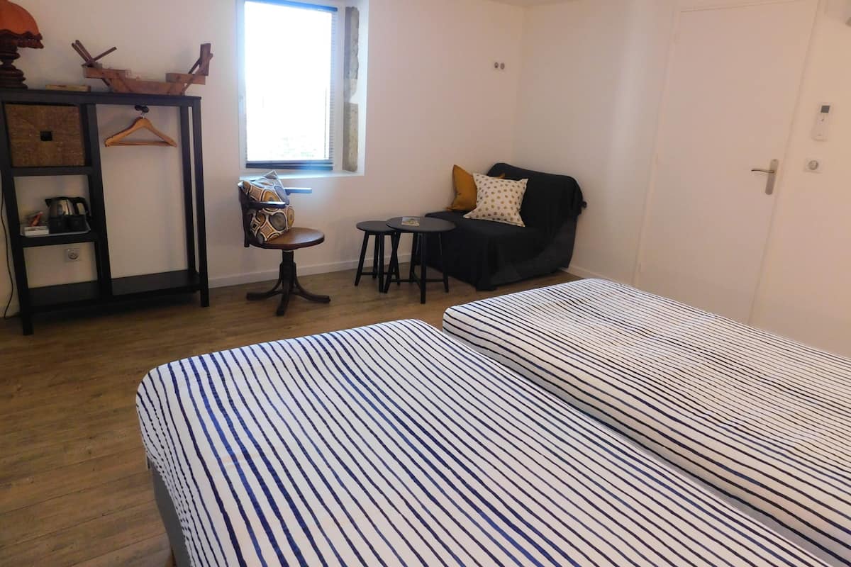 A bedroom with two beds, a small table, a chair, a shelf with items, and a window.