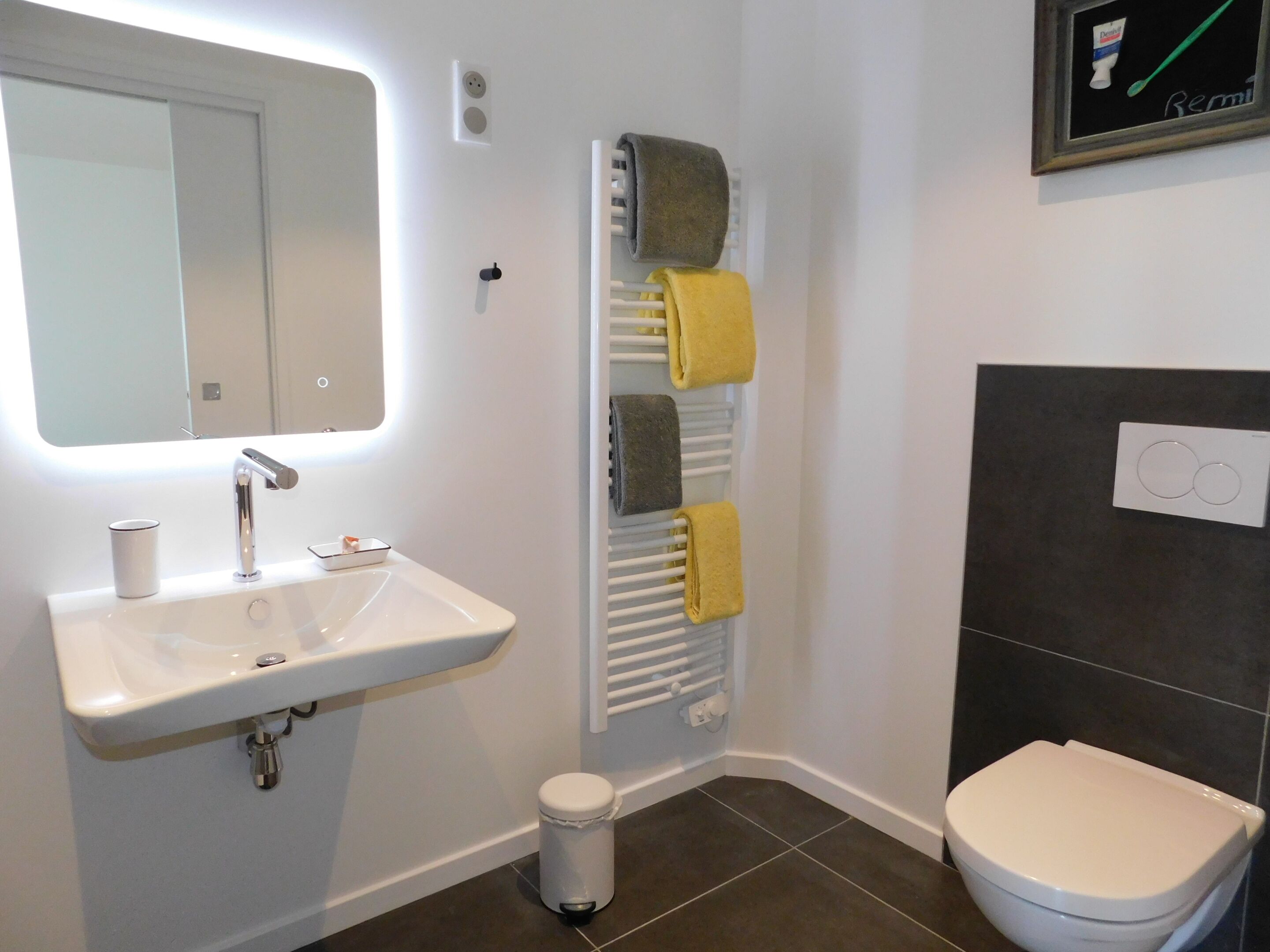 A bathroom with a white sink, a mirror, a towel rack with towels, a toilet, and a small trash bin.