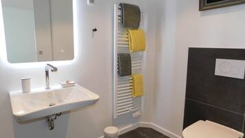 A bathroom with a white sink, a mirror, a towel rack with towels, a toilet, and a small trash bin.
