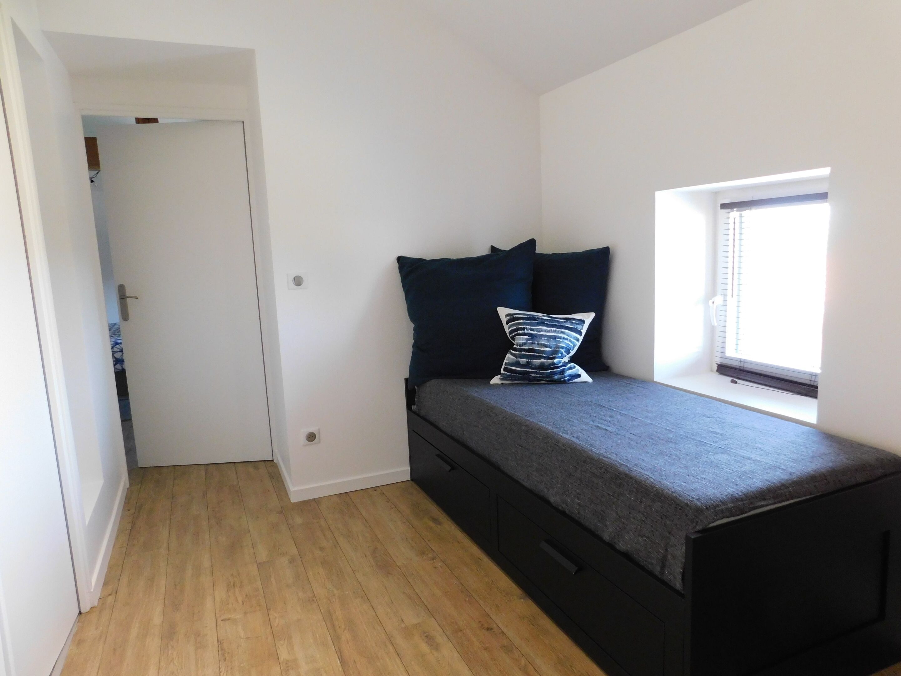 A small, compact room with a bed, a window, and a door leading to another room.