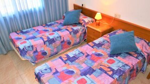 Dos camas individuales con ropa de cama colorida, una mesita de noche con lámpara y una ventana con cortinas.