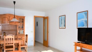 Una sala de estar con una mesa de comedor de madera, sillas, un televisor sobre un mueble de madera, y dos cuadros en la pared.