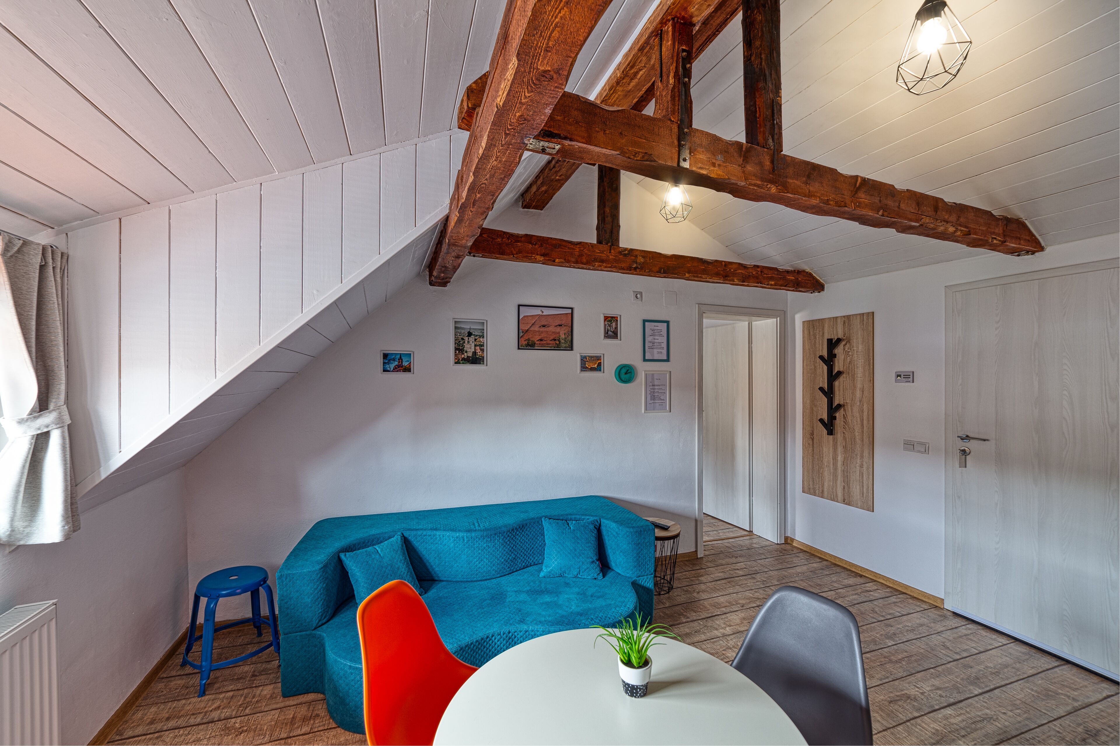 A room with a sloped ceiling, wooden beams, a blue sofa, a red chair, a small table with a plant, and a door with a wooden panel.
