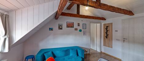 A room with a sloped ceiling, wooden beams, a blue sofa, a red chair, a small table with a plant, and a door with a wooden panel.