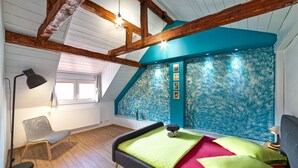 A bedroom with a sloped ceiling, wooden beams, a bed with green and red bedding, a chair, and a wall with a blue floral pattern. - T House (Sibiu)