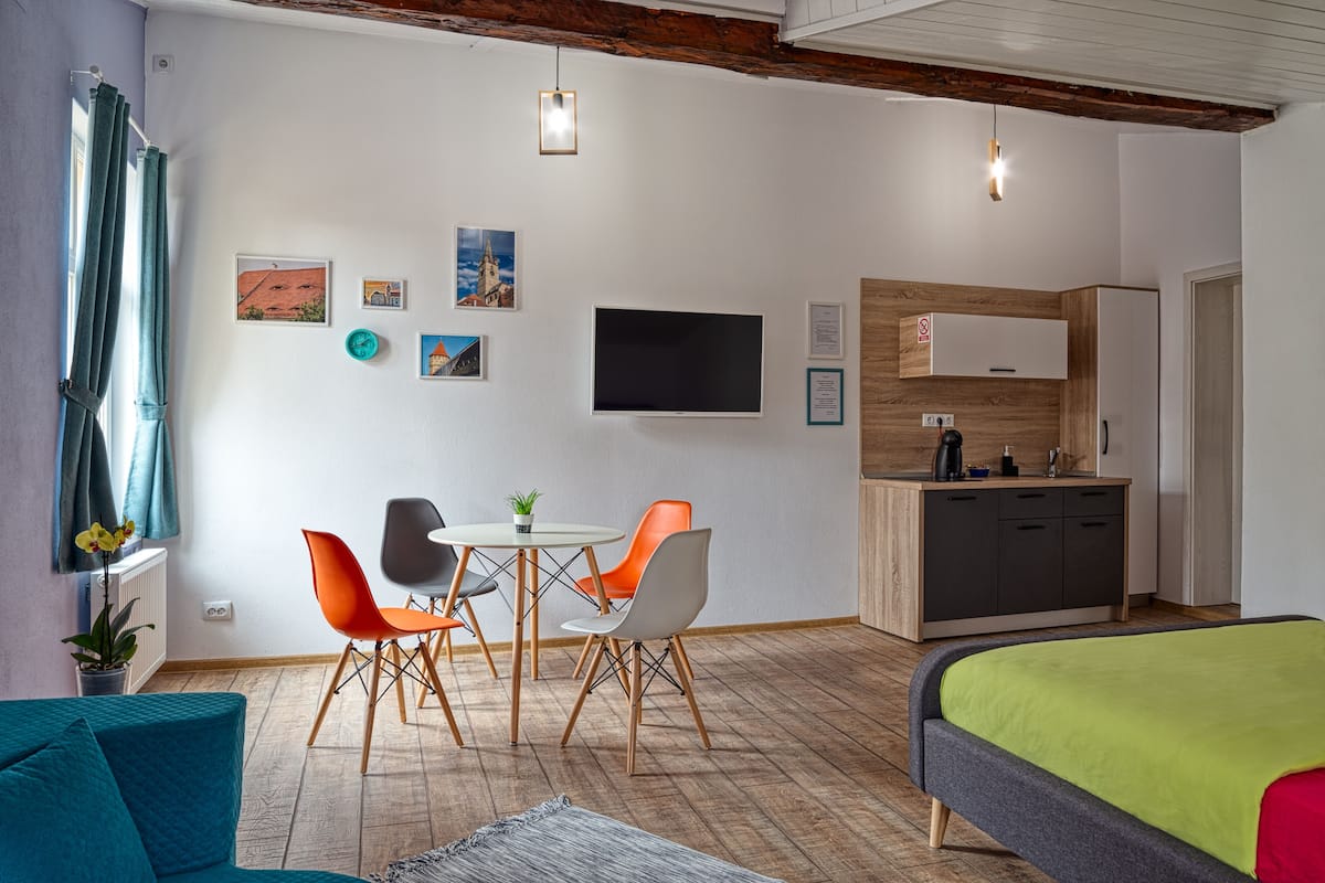 A modern room with a bed, a dining table with chairs, a TV, and a kitchenette.