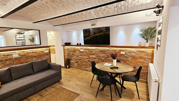 A cozy room with a gray sofa, a round wooden table with chairs, a brick accent wall, and a large mirror. - T House (Sibiu)