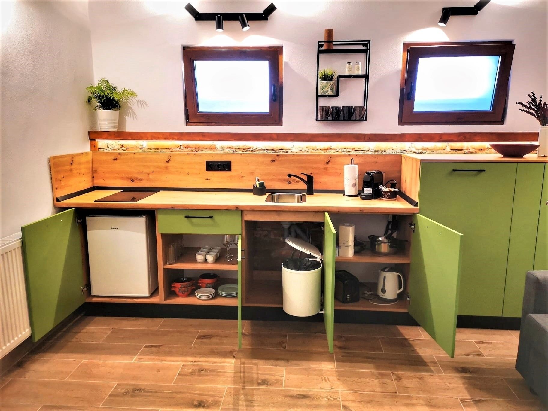 A compact kitchen with a wooden countertop, green cabinets, a small refrigerator, and two windows.