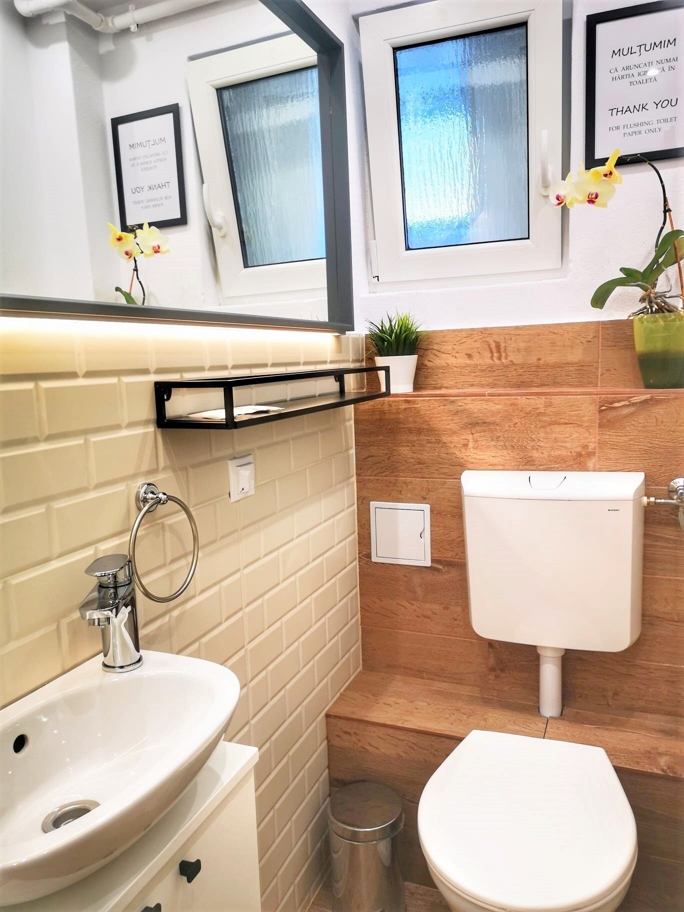 Ein modernes Badezimmer mit einem weißen Waschbecken, einer Toilette und einem Spiegel über dem Waschbecken. Zwei gerahmte Schilder an der Wand, eines bedankt sich für das Entsorgen von Toilettenpapier und das andere für die Nutzung der Toilette.