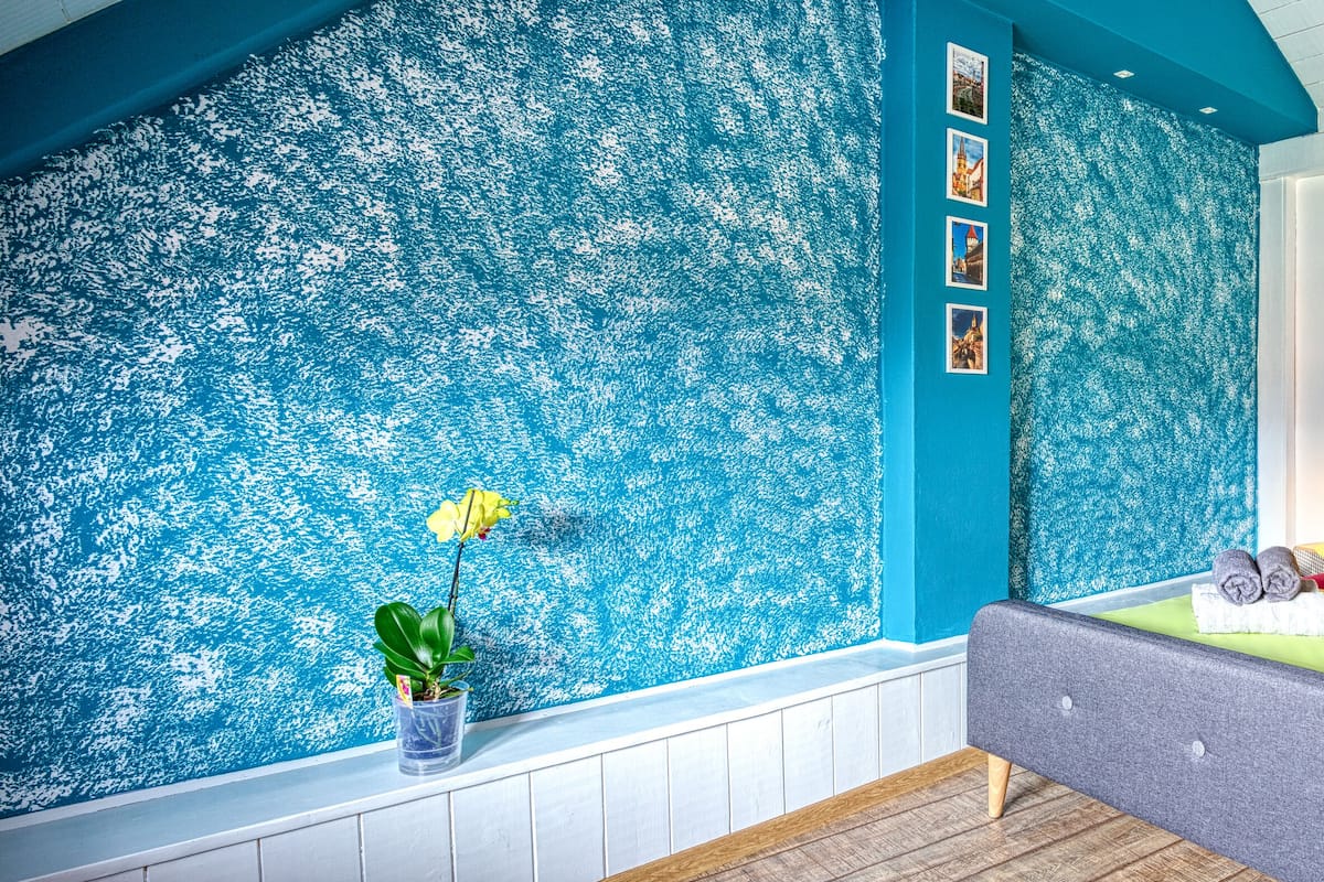 A bedroom with a blue textured wall, a grey bed with green and yellow pillows, a small potted plant, and a bedside lamp.