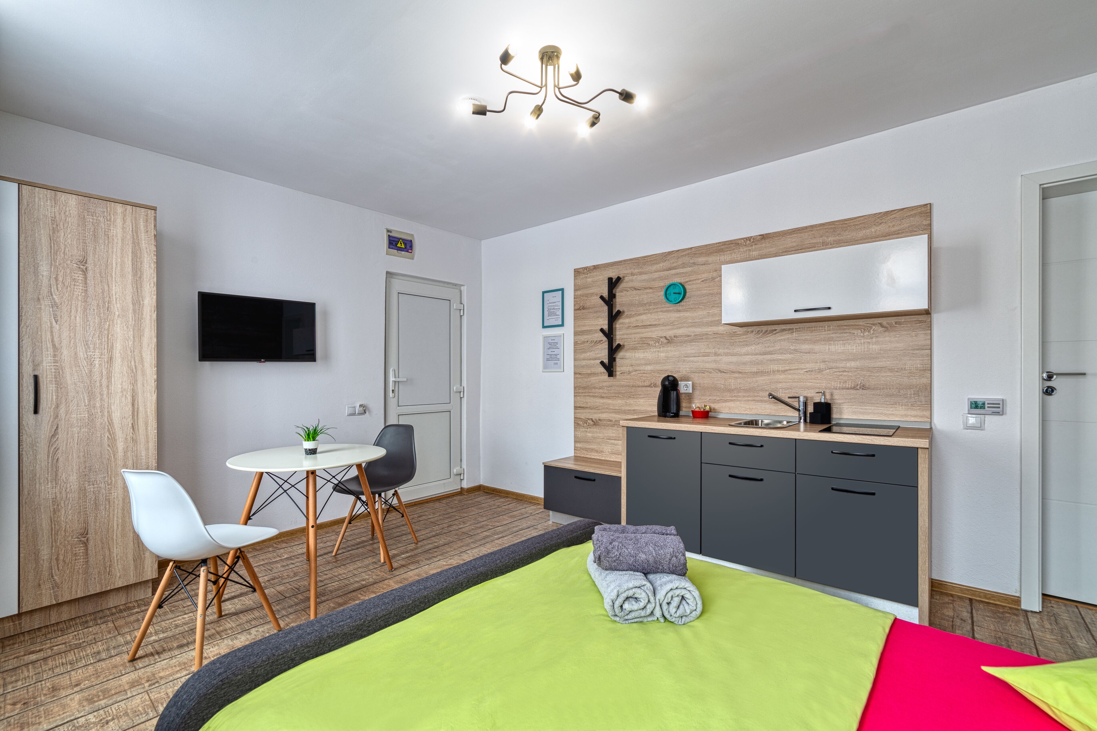 Una habitación de hotel moderna con un armario de madera, una zona de comedor con mesa y sillas, una kitchenette y una cama con manta verde, además de un televisor montado.