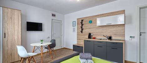 Una habitación de hotel moderna con un armario de madera, una zona de comedor con mesa y sillas, una kitchenette y una cama con manta verde, además de un televisor montado.